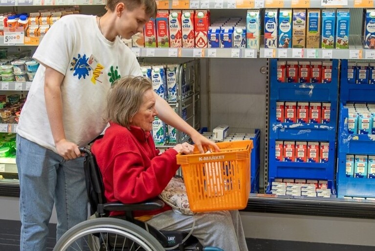 Maitohyllyn edessä ihminen pyörätuolissa ja toinen avustamassa.